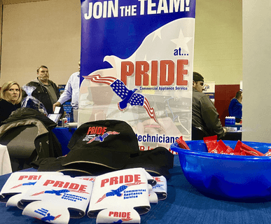 Two male technicians in safety gear are working on a commercial stainless steel conveyor system. One in a neon yellow shirt is using a tool that emits sparks, while the other in a similar shirt and a decorated hard hat, is looking down at the equipment.