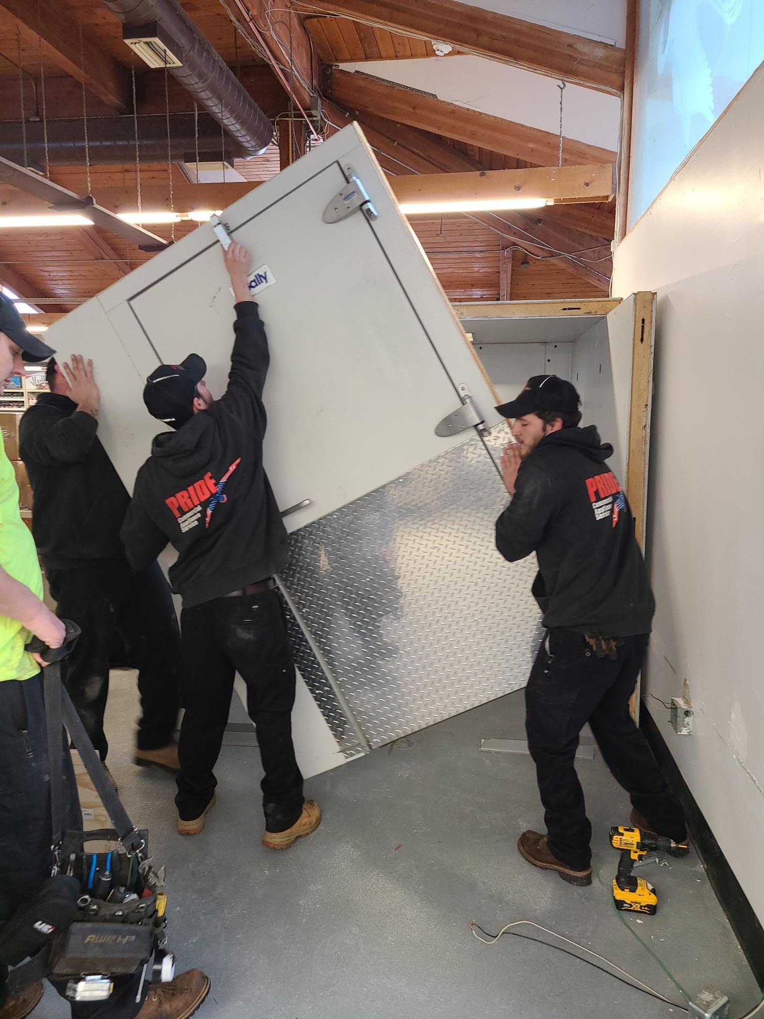 Three Pride Commercial Appliance Service workers are installing a large white walk-in cooler door with a diamond plate kick plate in a commercial space with a high wooden ceiling. Tools are visible at the bottom of the frame.