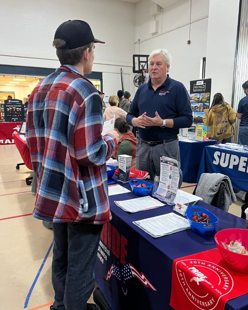 At a "PRIDE Commercial Appliance Service" booth, a PrideCAS manger speaks to a student about careers. Informational flyers fill the table between them.