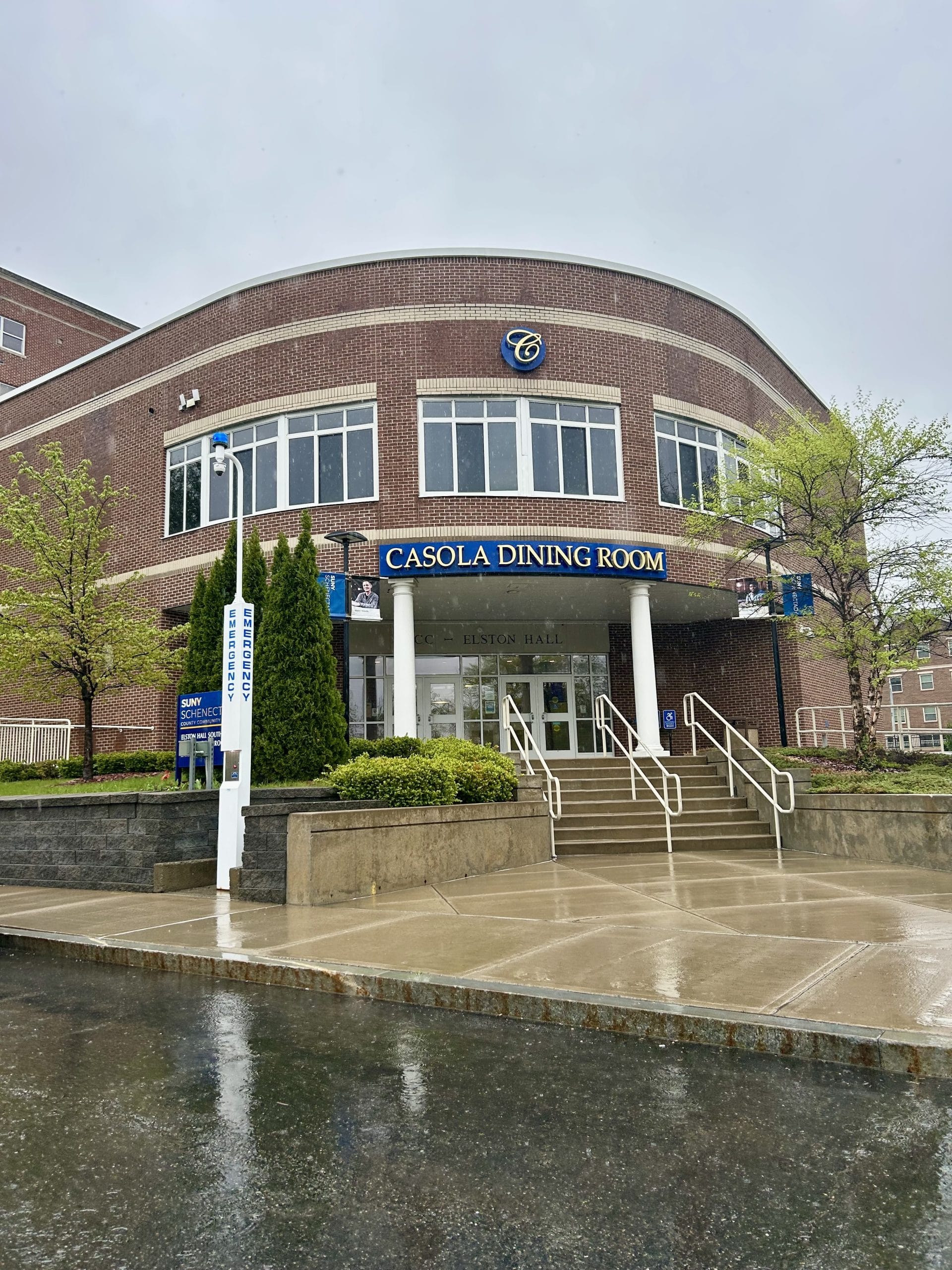 The Casola Dining Room, a curved brick building, features a prominent sign, steps to its entrance, and landscaping on a wet day.