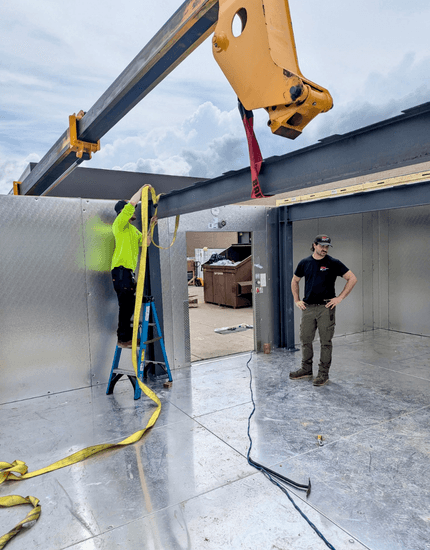 Two pridecas workers at the Central Valley, Utica, NY installation use a yellow lifting arm and strap inside a large metal-paneled room.