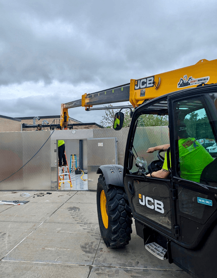A JCB telehandler, operated by a pridecas technician in a bright green shirt, is positioned near a large metal walk-in unit at a Central Valley, Utica, NY installation site. Another pridecas worker is on a ladder near the unit.