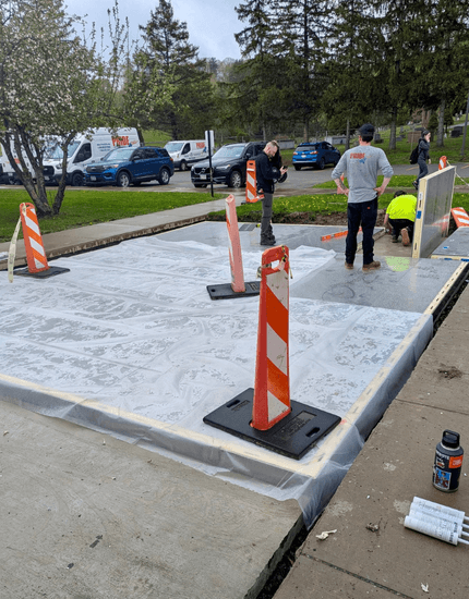 A new concrete slab covered with white plastic is surrounded by orange and white traffic cones at an installation site in Central Valley, Utica, NY. Several workers are present, and vehicles are parked in the background.