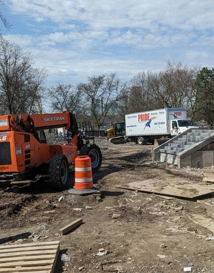 A construction site with an orange SKYTRAK telehandler, an orange traffic barrel, a white "PRIDECAS" service truck, and a concrete staircase under a partly cloudy sky.