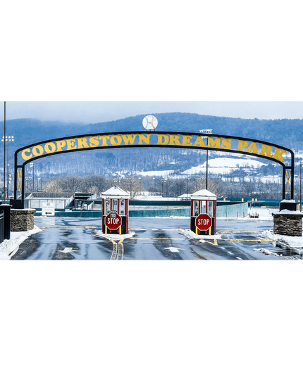 The entrance to Cooperstown Dreams Park in winter, with a large arched sign bearing its name in yellow letters, two stop booths, and snow-covered fields and mountains in the background.