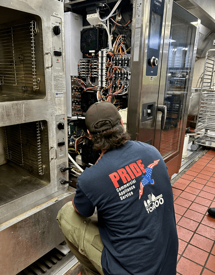 "Pride Commercial Appliance Service technician in branded uniform servicing electrical control panel of commercial kitchen equipment in restaurant setting with tile floor"