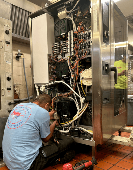 Pride technician in company uniform working on internal components of opened commercial kitchen equipment, with complex wiring and control systems visible in professional kitchen setting.