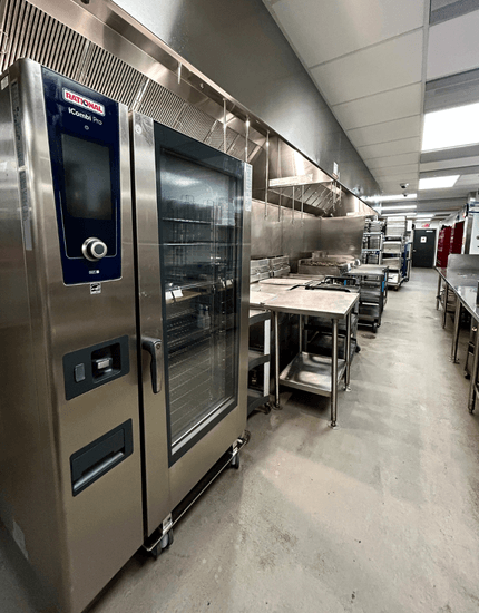 Large commercial kitchen with Rational combi oven, stainless steel ventilation hood, work tables, and equipment storage in institutional food service facility