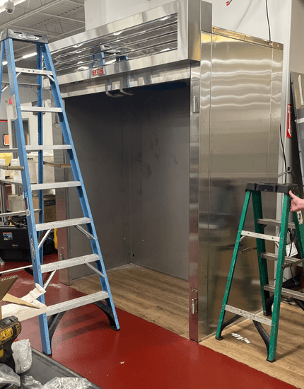 Large stainless steel commercial walk-in cooler or freezer unit being installed in restaurant kitchen, with blue and green step ladders positioned nearby during construction