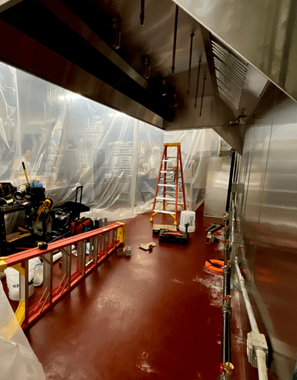 "Commercial kitchen under construction with red step ladder, plastic sheeting covering equipment, stainless steel ventilation hood, and red epoxy floor coating