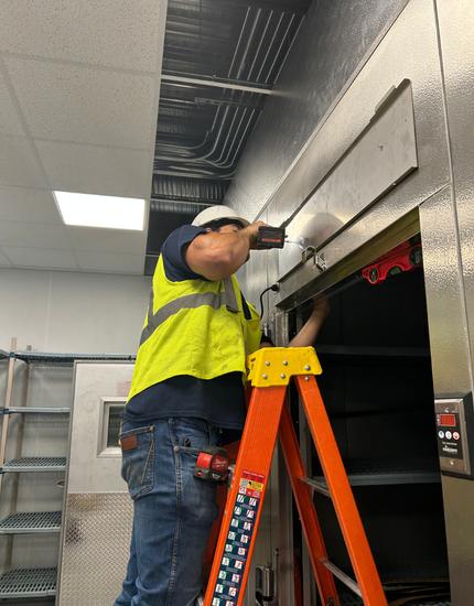 PrideCAS technician stands on ladder using drill to install an AC unit in walk in refrigerator